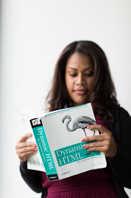 A woman with dark, shoulder-length hair is holding a book titled 'Dynamic HTML' with a flamingo illustration on the cover. She is dressed in a dark blazer and a burgundy top, standing indoors with a p