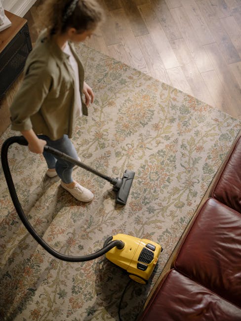 A woman using a yellow vacuum cleaner to perform surface cleaning on a patterned, beige and pastel-colored rug in a living room. She is dressed casually in a beige jacket, white sneakers, and jeans. T